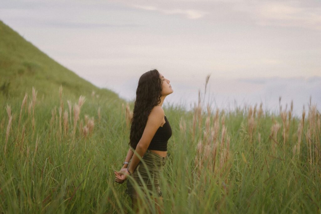 Peaceful woman standing in tall grass with closed eyes, doing breathing exercises for stress and inner calm.