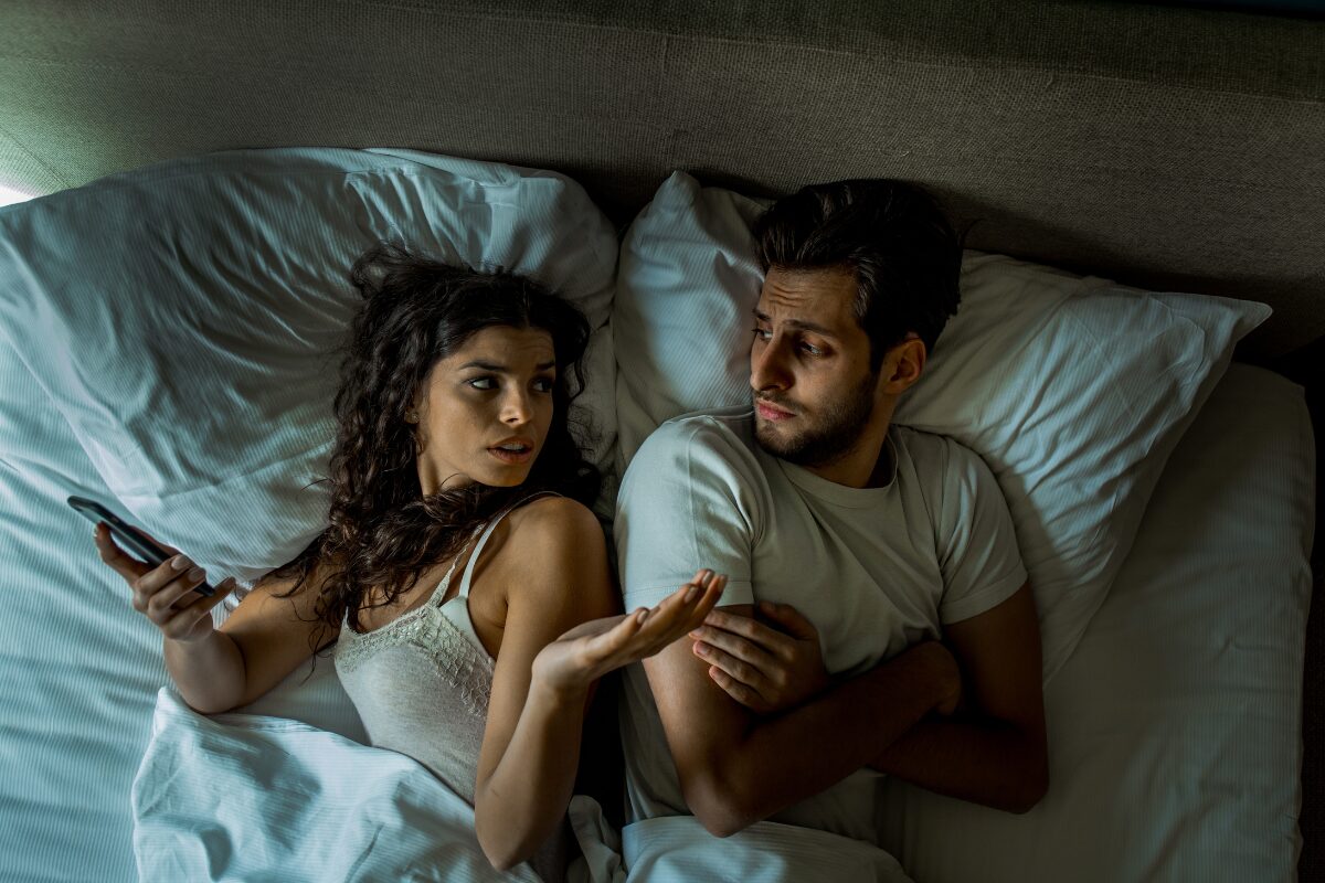 Woman checking her phone while man looks back in bed – how to stop being jealous in a relationship