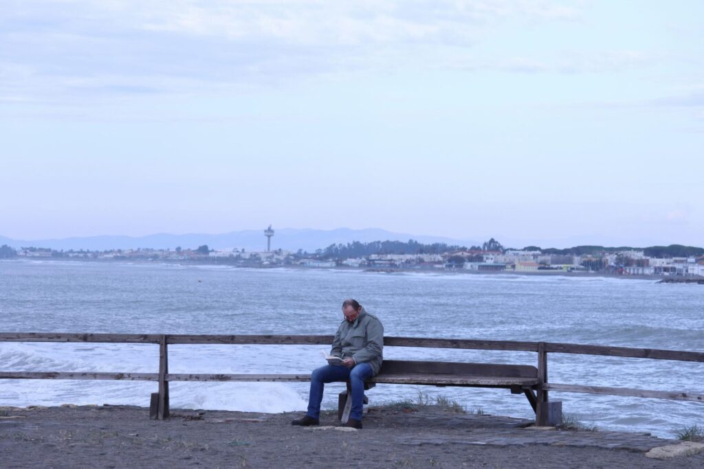 Older man sitting alone on a bench by the sea in cold weather, reading a book, showing how to cope with loneliness when you have no one.