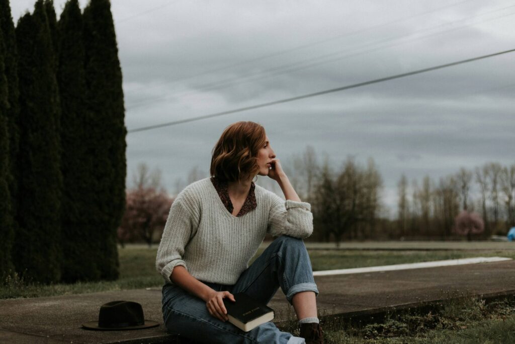 Woman sitting on a sidewalk, holding her head with one hand and a closed book in the other – illustrating mental exhaustion and the inner consequences of self-criticism.