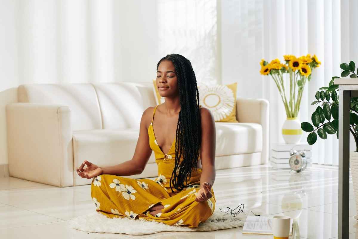 How to start meditating at home as a beginner: dark-skinned woman sitting on the floor in a yellow outfit, practicing meditation in her home