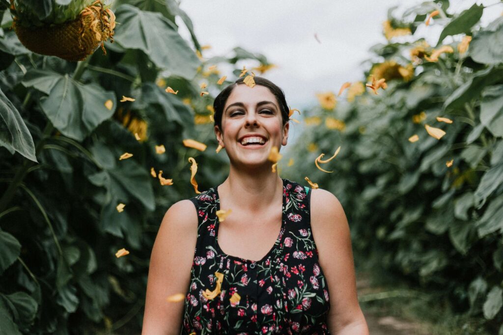 A joyful woman standing among sunflowers with petals falling on her — radiating happiness and showing clear signs you truly love yourself.