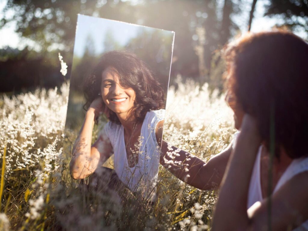 A smiling woman looking at herself in a mirror while standing in a field — capturing deep self-acceptance and signs you truly love yourself.

