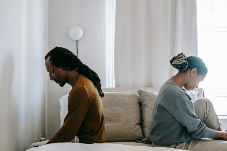 "Couple sitting on opposite sides of the bed, turned away from each other, illustrating one of the common mistakes during partner disagreements—emotional withdrawal and lack of communication.
