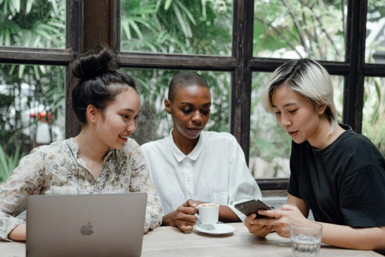 Three female friends sitting at a table, looking at a phone and laptop while working together — how to know if your friend is fake moments might hide behind everyday interactions like these.