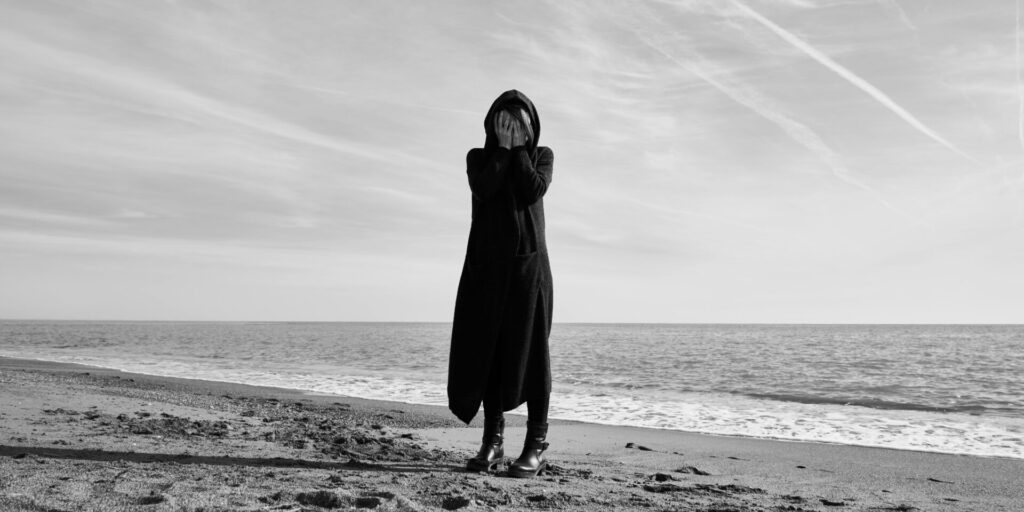 Black and white image of a lonely woman standing on the beach, covering her face, feeling isolated while learning how to deal with loneliness after a breakup.