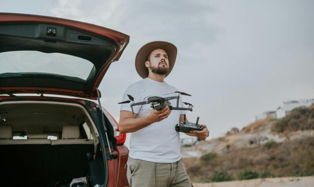 Man standing by his car in nature holding a drone, wearing a hat, symbolizing reasons why you don't achieve goals
