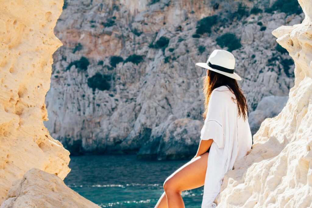 Woman leaning against a rock and gazing out at the sea from above, enjoying peaceful and reflective summer self-care activities.