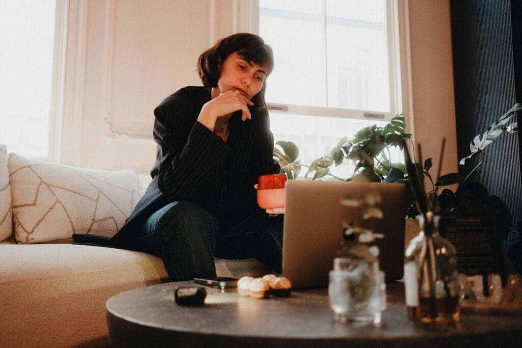 Woman sitting on a couch, holding a cup and looking thoughtfully at her laptop, focused on learning how to stop worrying about past mistakes and feel free again.

