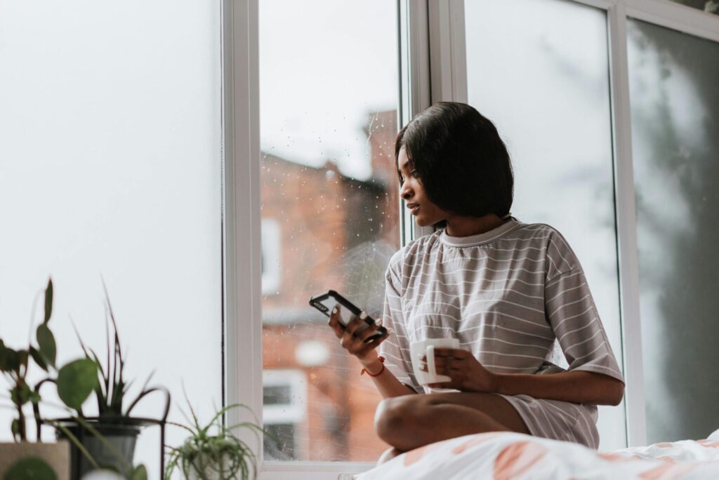 Woman sitting by the window on a rainy day, holding a phone and a cup, enjoying cozy rainy day activities for adults who hate being bored.
