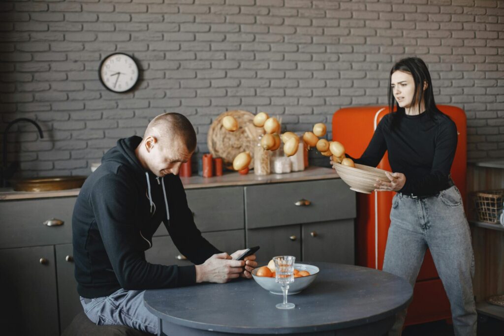 A woman is angry in the kitchen at her partner, who is scrolling on his phone.
