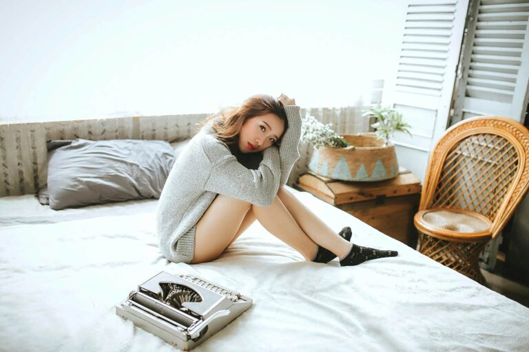 Young woman sitting alone on a bed with a typewriter beside her, looking at the camera — a quiet moment that reflects self-care ideas for highly sensitive people.