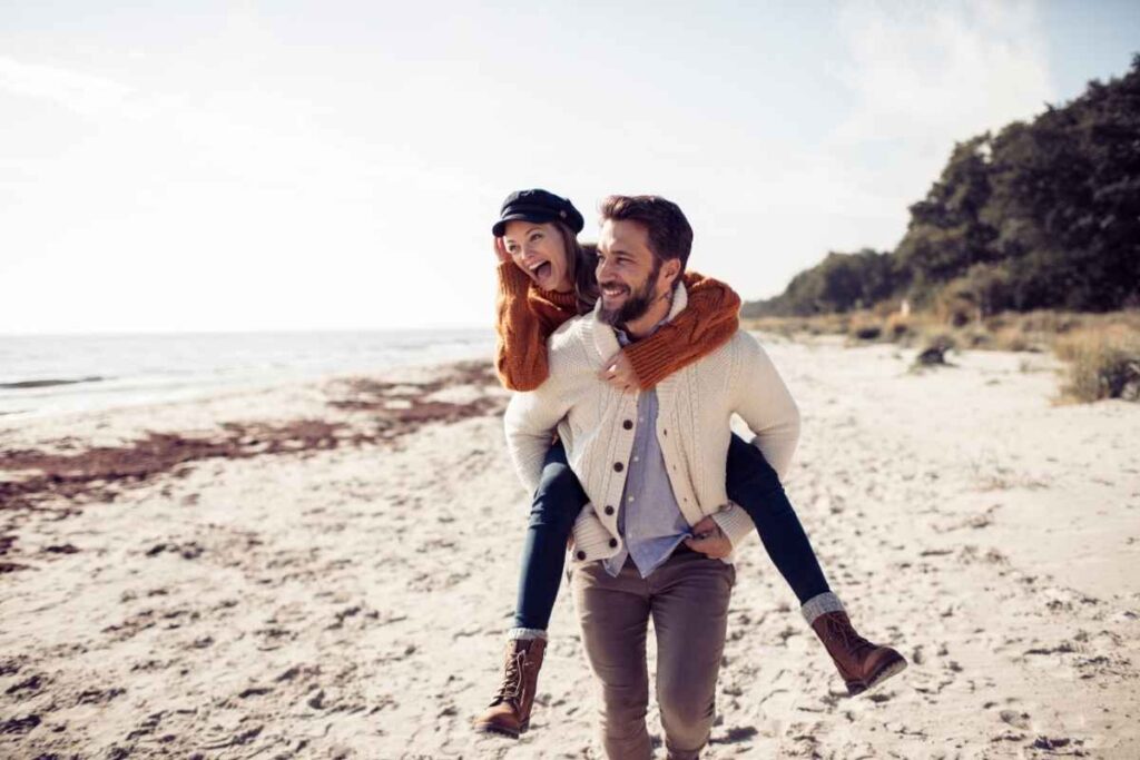 a playful couple at the beach, with the man carrying the woman on his back, representing excitement and the early honeymoon phase