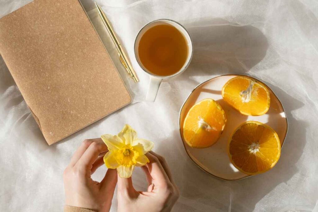 Spring journal prompts: cozy spring journaling scene with a journal, tea, mandarin, and hands holding daffodils, inspiring mindful reflection and goal setting