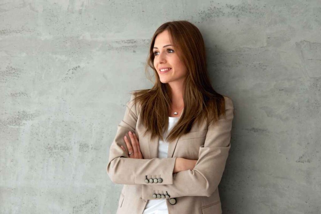 Habits that destroy your confidence: a confident woman in business attire standing against a wall, showing strength and empowerment despite common confidence-diminishing habits
